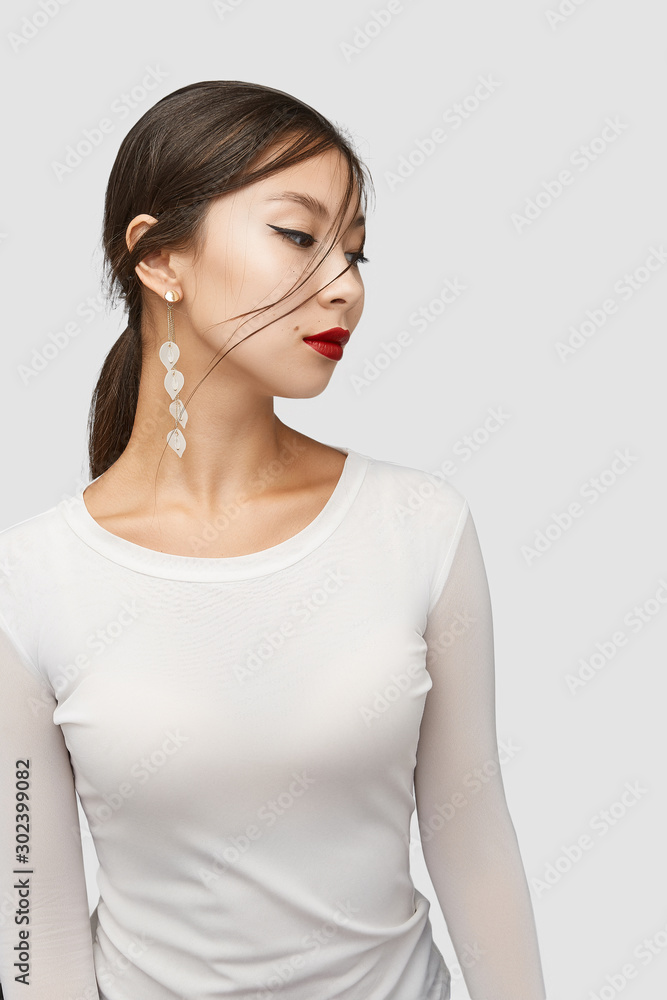Medium shot of a young Asian woman in an ivory white blouse and with a stud earring made as a golden disc with dangling string of white gem leaf-shaped pendants and chains with crystals. Medium shot of a young Asian woman in an ivory white blouse and with a stud earring made as a golden disc with dangling string of white gem leaf-shaped pendants and chains with crystals.