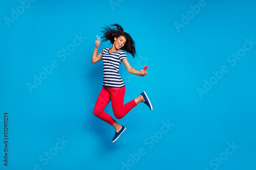 Fototapeta Naklejka Na Ścianę i Meble -  Turned full length body size photo of cheerful positive excited crazy girl filming herself taking selfie wearing trousers pants red striped t-shirt jumping isolated vibrant color blue background