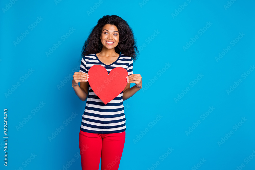 Portrait of positive cheerful afro american girl hold paper card big red heart present from romantic boyfriend on 14-february wear striped t-shirt casual style clothing isolated blue color background Portrait of positive cheerful afro american girl hold paper card big red heart present from romantic boyfriend on 14-february wear striped t-shirt casual style clothing isolated blue color background