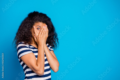 Photo of scared frightful girlfriend hiding her face to avoid negative emotions wearing striped t-shirt looking through hand near empty space isolated vivid blue color background photo