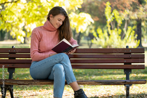 Young woman sitting on a bench in autumn park and is reading a book.