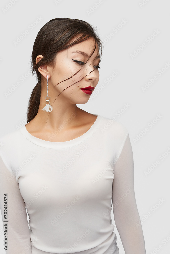 Medium shot of a young Asian woman in an ivory white blouse and with a stud earring made as a sparkling crystal string with a gem flower on silver rings and golden cylinder.