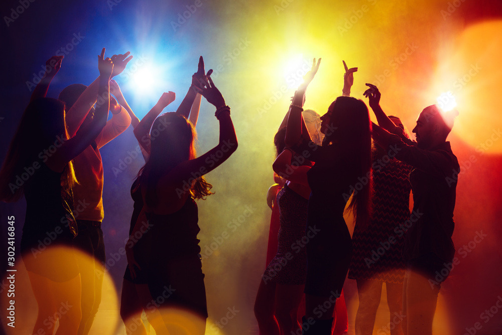 Fototapeta Freedom. A crowd of people in silhouette raises their hands on dancefloor on neon light background. Night life, club, music, dance, motion, youth. Yellow-blue colors and moving girls and boys.