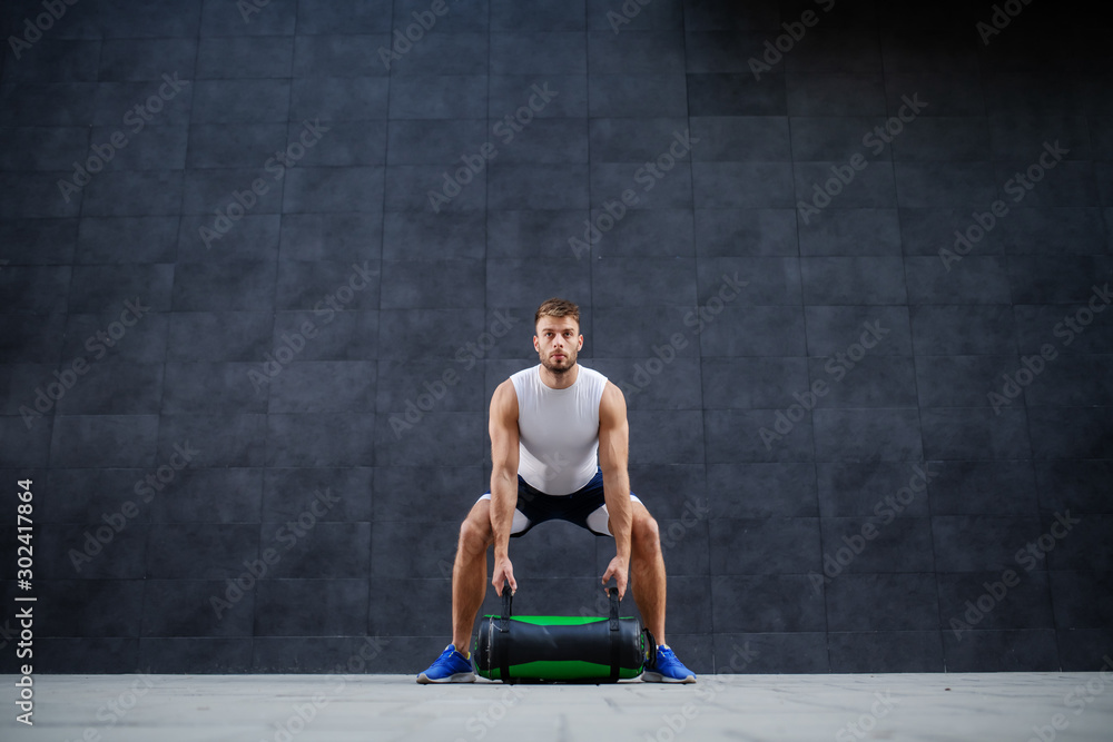 Handsome muscular bearded caucasian man in shorts and t-shirt lifting training bag while standing outdoor in front of gray wall. Handsome muscular bearded caucasian man in shorts and t-shirt lifting training bag while standing outdoor in front of gray wall.