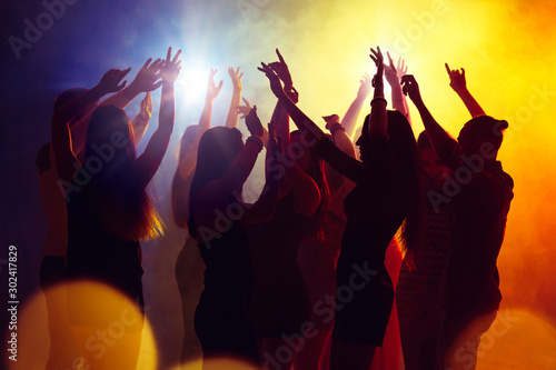 Young. A crowd of people in silhouette raises their hands on dancefloor on neon light background. Night life, club, music, dance, motion, youth. Yellow-blue colors and moving girls and boys.