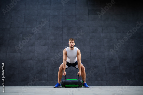 Handsome muscular bearded caucasian man in shorts and t-shirt lifting training bag while standing outdoor in front of gray wall.