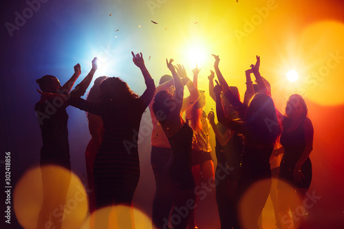 Confetti. A crowd of people in silhouette raises their hands on dancefloor on neon light background. Night life, club, music, dance, motion, youth. Yellow-blue colors and moving girls and boys.