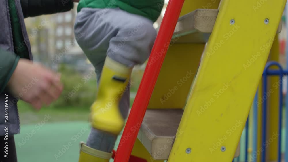 Medium tracking shot of young Caucasian boy in warm jacket, rubber boots and hat climbing up ladder at playground, and hands of unrecognizable father who is standing behind and guarding him Medium tracking shot of young Caucasian boy in warm jacket, rubber boots and hat climbing up ladder at playground, and hands of unrecognizable father who is standing behind and guarding him