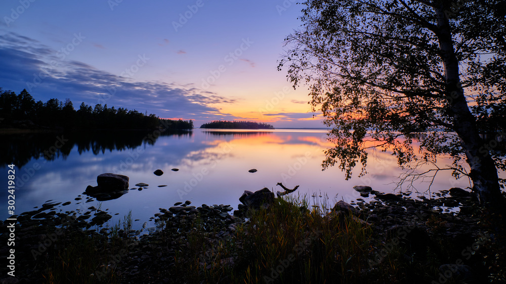 Evening by a lake with trees and beach in the foreground and sunset in the background