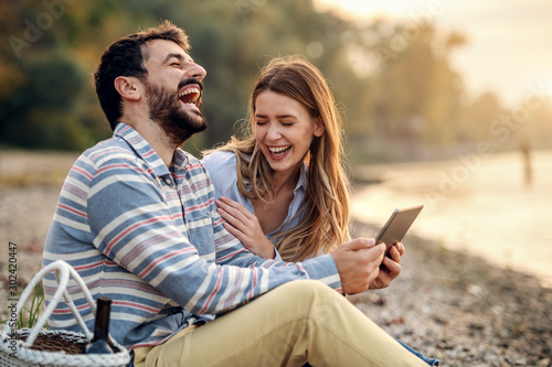 happy laughing caucasian fashionable young couple sitting on coast near river and using tablet. Man holding tablet. Next to man is picnic basket.