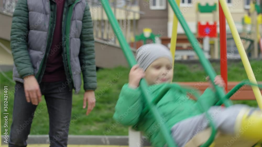 Medium shot of delighted 4-year-old Caucasian boy in warm jacket, rubber boots and hat riding on swing in playground, and active young father pushing him Medium shot of delighted 4-year-old Caucasian boy in warm jacket, rubber boots and hat riding on swing in playground, and active young father pushing him