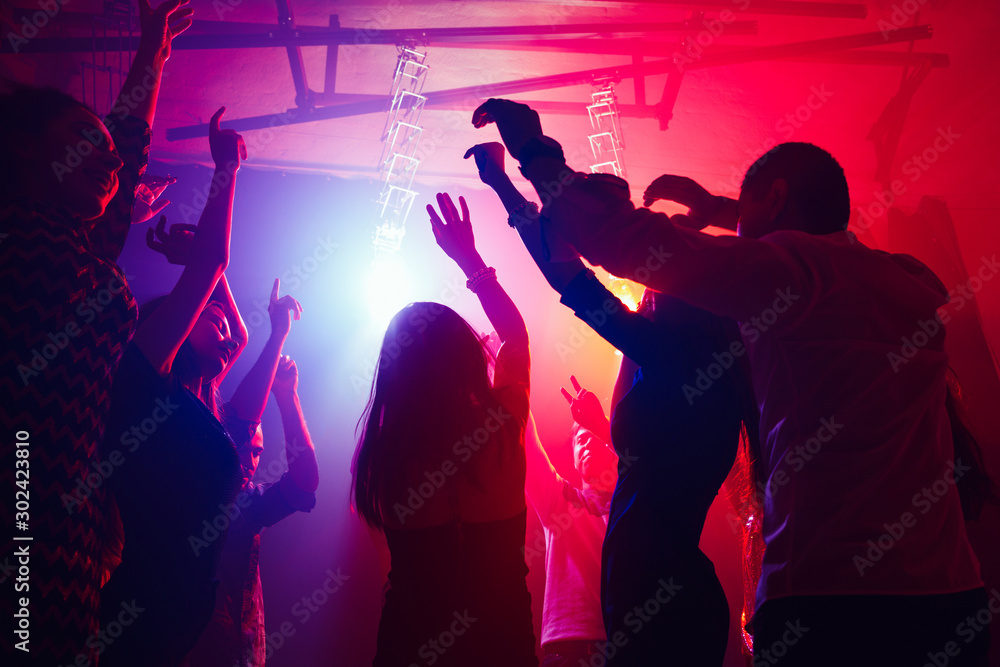 Cheers. A crowd of people in silhouette raises their hands on dancefloor on neon light background. Night life, club, music, dance, motion, youth. Purple-pink colors and moving girls and boys.