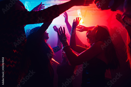 A crowd of people in silhouette raises their hands on dancefloor on neon light background. Night life, club, music, dance, motion, youth. Purple-pink colors and moving girls and boys.