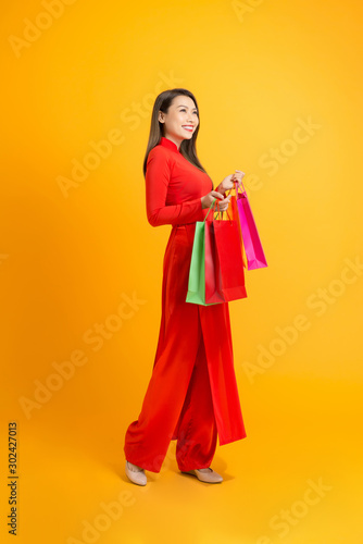 Beautiful Asian girl holding colorful shopping bags while wearing Ao Dai Vietnam over yellow background.