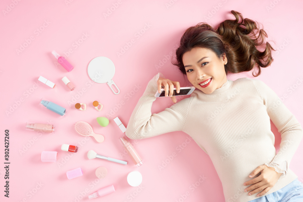 Young Asian beautiful woman show smart phone with her cosmetic makeup tools - she is lying on the pink floor. Top view Young Asian beautiful woman show smart phone with her cosmetic makeup tools - she is lying on the pink floor. Top view