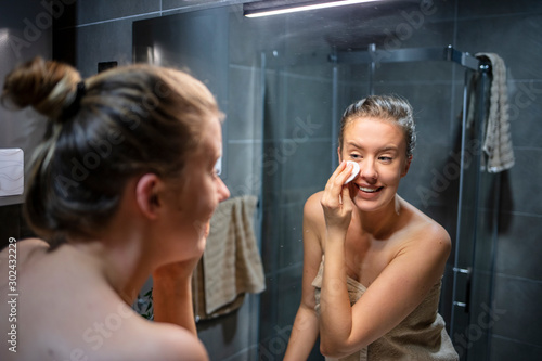 Young woman in bathrobe looking in bathroom mirror. Portrait of beautiful girl in bathrobe and with towel cleaning her face with sponge and smiling, standing in bathoom.