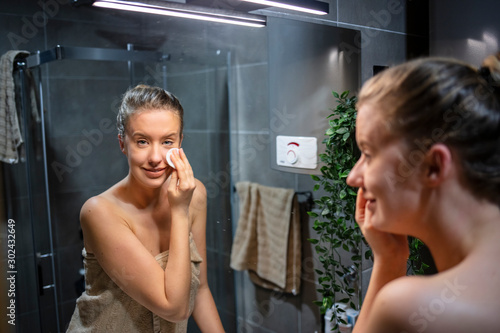 Beautiful brunette woman removing makeup from her face. Taking good care of her face. Beautiful young woman cleaning her face with sponge and smiling while standing against a mirror in bathroom