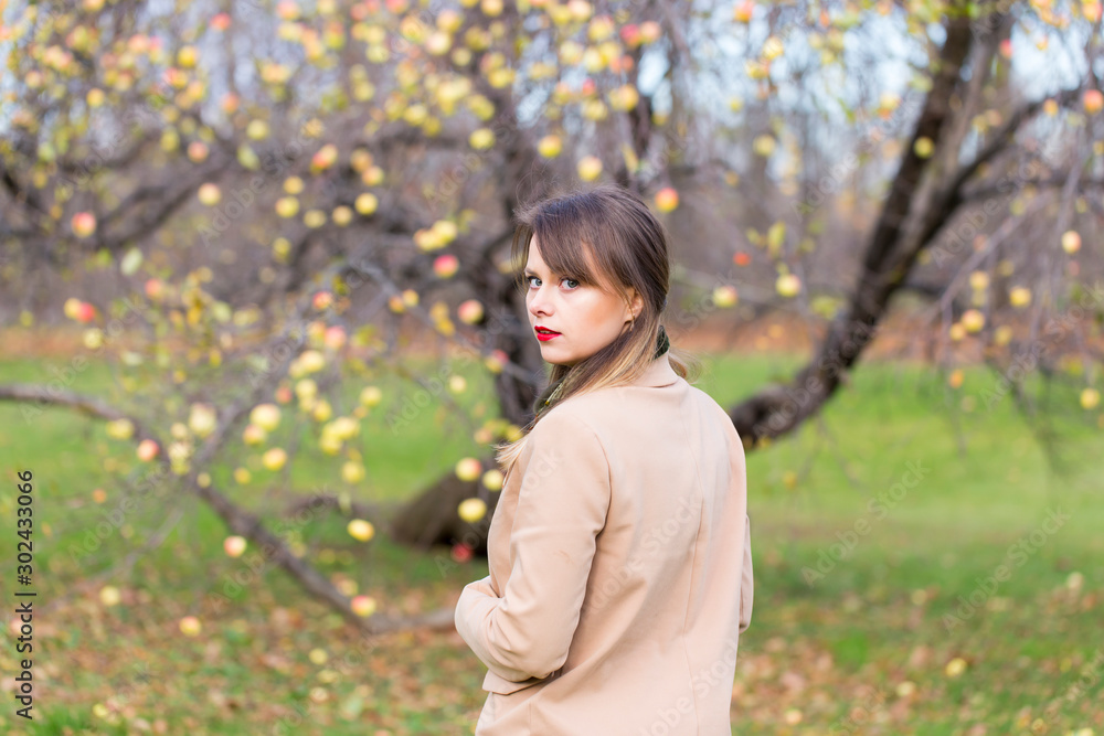Pretty young woman with long straight hair wearing beige coat standing in front of apple tree in late fall turning back and looking over her left shoulder Pretty young woman with long straight hair wearing beige coat standing in front of apple tree in late fall turning back and looking over her left shoulder