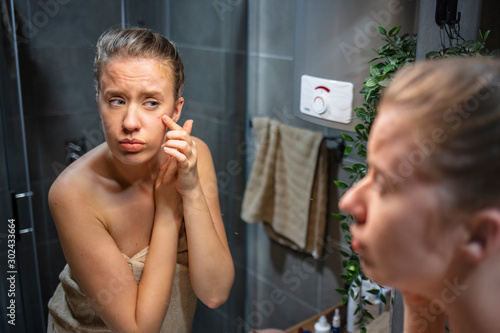 Girl having problems with skin looking in the mirror. Portrait of beautiful young woman squeezing pimples while looking at the mirror. Woman has problems with skin on the face