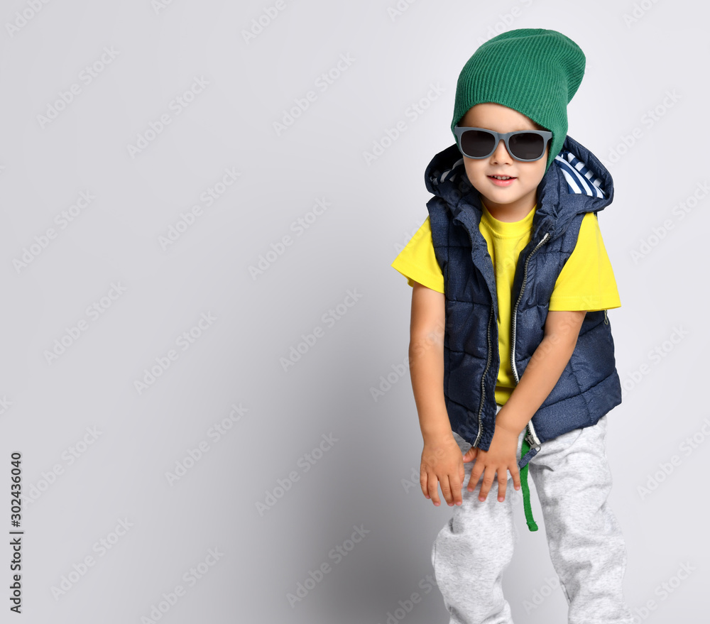 Stylish cheerful little boy 4 years old in sweatpants, a green hat, glasses and a down vest has fun Stylish cheerful little boy 4 years old in sweatpants, a green hat, glasses and a down vest has fun