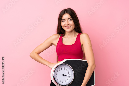 Pretty young girl with weighing machine over isolated pink background with arms at hip and holding weighing machine