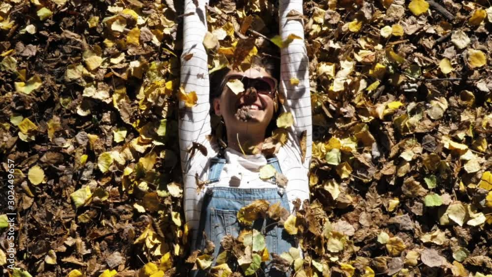 Portrait of cheerful young woman in denim overall is lying on autumnal yellow leaves, looking at camera, smiling and throwing up a leaves. Happy fall season concept.