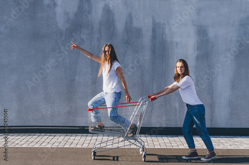Two happy hipster girls staying on shopping cart outdoors