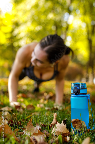 blue bottle of water on green grass. sporty woman doing outdoor workout in park on background. focus on bottle