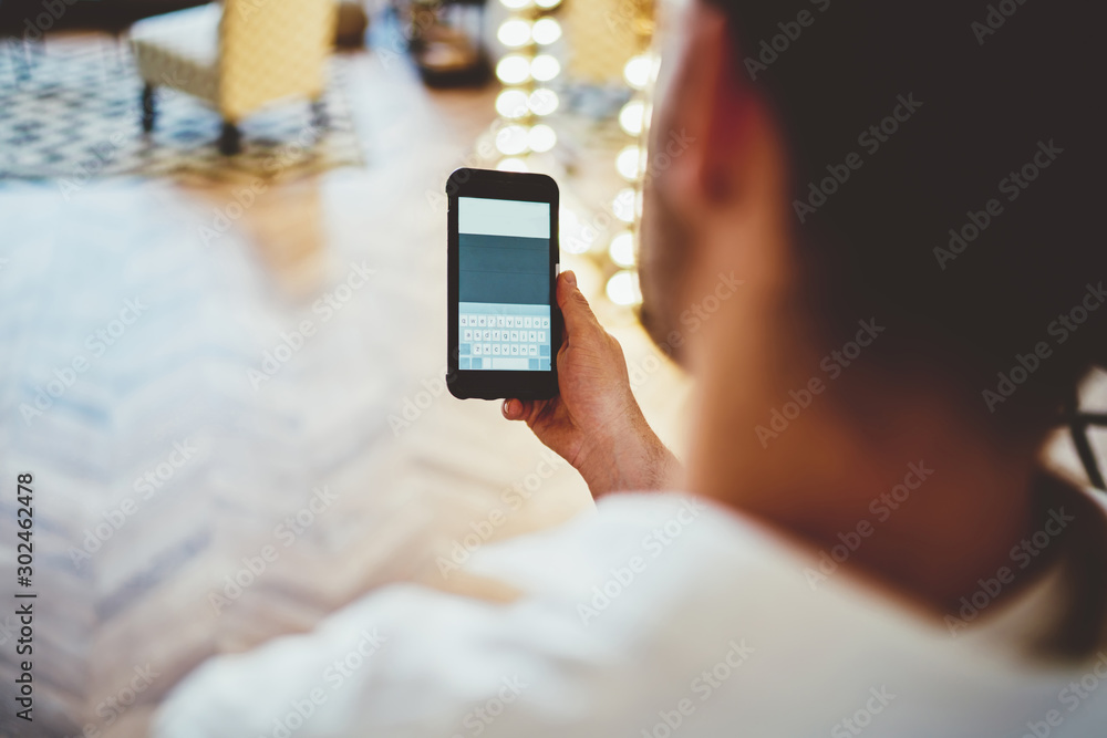Back view of millennial male using mobile phone for chatting in social networks via app, cropped image of hipster guy holding smartphone while reading information from web page at home interior. Back view of millennial male using mobile phone for chatting in social networks via app, cropped image of hipster guy holding smartphone while reading information from web page at home interior.