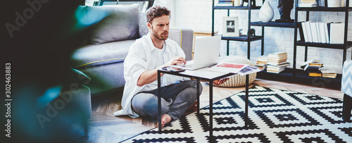 Serious caucasian millennial male making online booking via laptop computer sitting in modern interior living room, concentrated man freelancer concentrated on remote job using netbook at home.