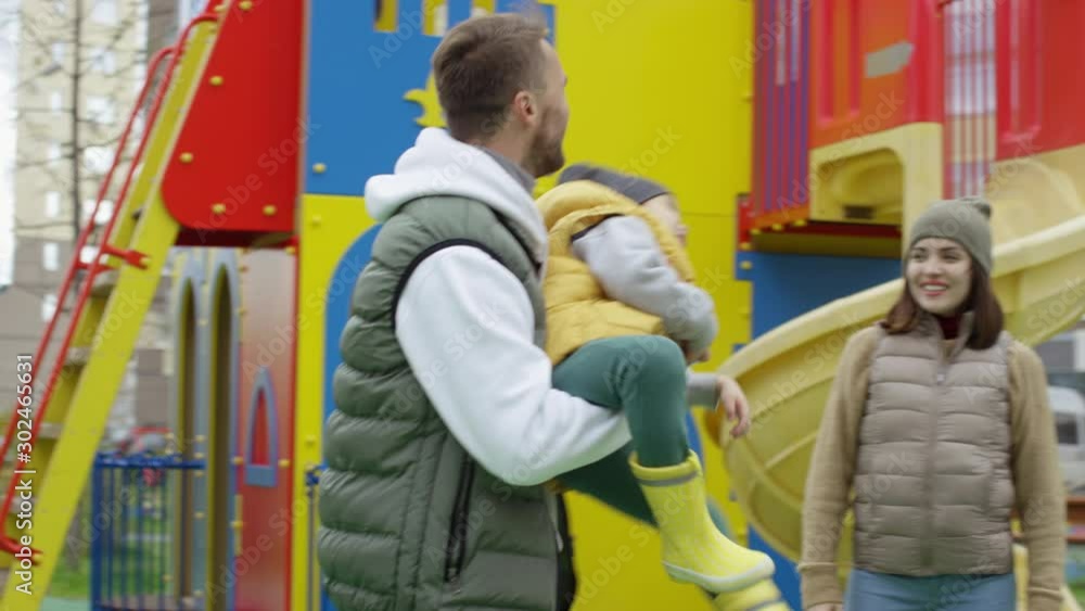 Medium tracking shot of warmly dressed laughing young Caucasian father having fun with son at outdoor playground, holding him in arms, spinning and running around cheerful mother Medium tracking shot of warmly dressed laughing young Caucasian father having fun with son at outdoor playground, holding him in arms, spinning and running around cheerful mother