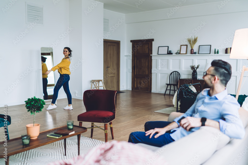 Man sitting on sofa and looking at woman standing in front of mirror in large room Man sitting on sofa and looking at woman standing in front of mirror in large room