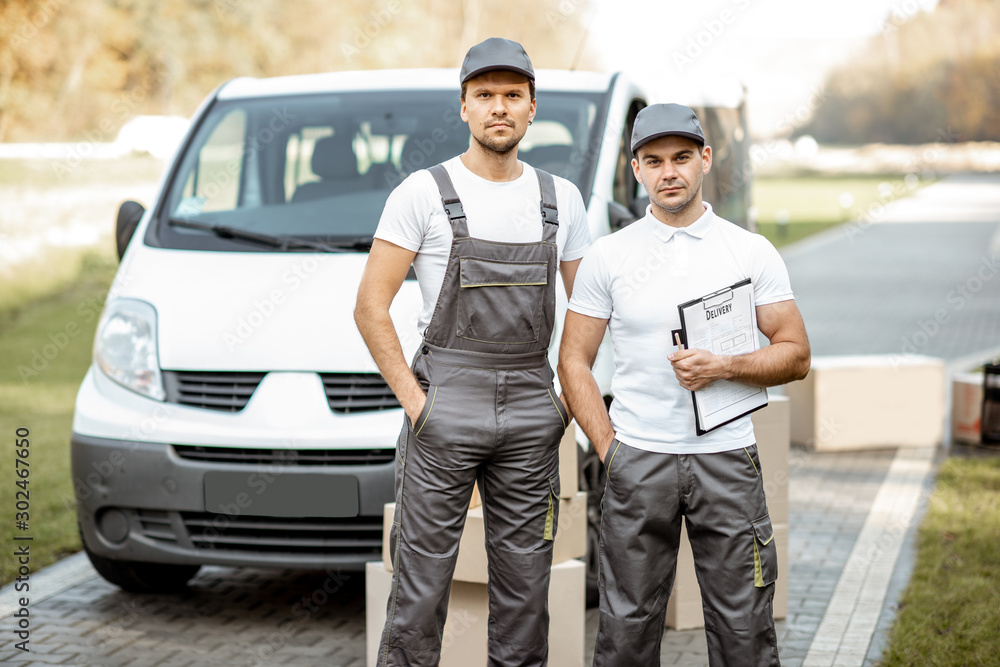Portrait of a two delivery men in uniform standing together with check list and cardboard boxes near the cargo van vehicle outdoors Portrait of a two delivery men in uniform standing together with check list and cardboard boxes near the cargo van vehicle outdoors