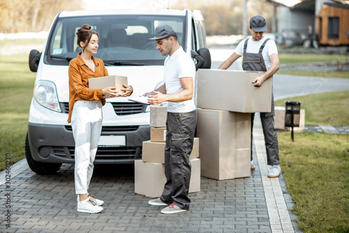 Courier with checklist delivering goods to a young woman by cargo van vehicle, mover with cardboard parcels on the background