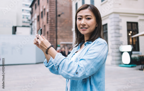 Portrait positive Asian woman in denim shirt smiling at camera while shooting online video and sharing to social networks via mobile application, happy tourist enjoying getaway in city historic center © BullRun