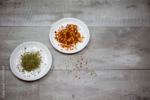 Dried red pepper flakes and dried basil in white dishes on a wooden background