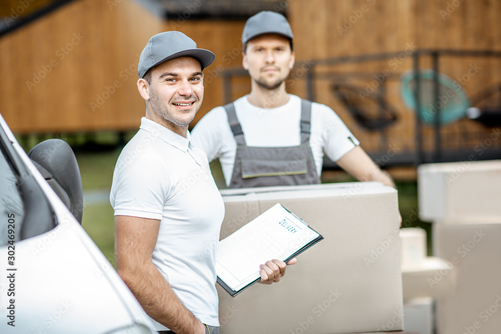 Portrait of a two delivery men in uniform standing together with check list and cardboard boxes near the cargo van vehicle outdoors Portrait of a two delivery men in uniform standing together with check list and cardboard boxes near the cargo van vehicle outdoors