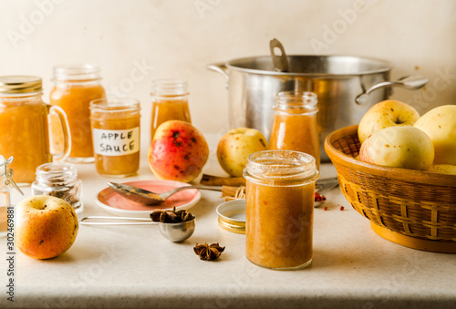 Fototapeta Naklejka Na Ścianę i Meble -  Freshly cooked homemade applesauce in glass jars, ingredients, plate, utensils on kitchen table, closeup view