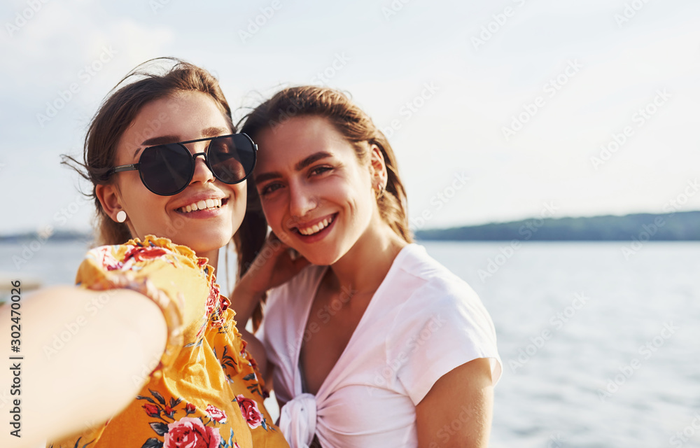 Selfie of two smiling girls outdoors that have a good weekend together at sunny day against the lake Selfie of two smiling girls outdoors that have a good weekend together at sunny day against the lake
