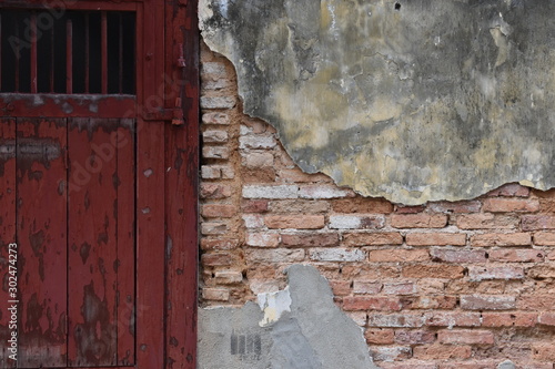 The red wooden door on the broken old concrete wall reveals a red brick inside.