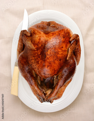 A Thanksgiving Turkey and carving knife on a white oval platter on an off-white table cloth. High angle in vertical format.