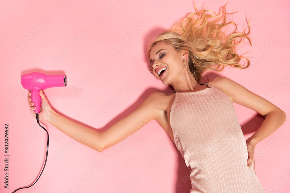 happy beautiful blonde woman lying and holding hairdryer isolated on pink happy beautiful blonde woman lying and holding hairdryer isolated on pink