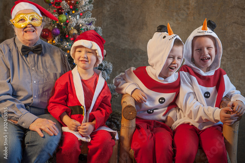 Happy brothers dressed in Christmas costumes of snowman. Attractive retired woman in bow tie and Santa Claus hat. Children grouped around grandmother in anticipation of Christmas presents. 