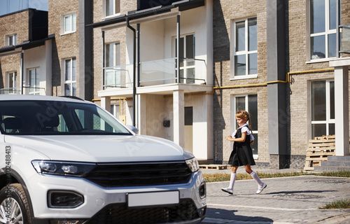 Little girl in school uniform runs ro a modern white car outdoors on the street