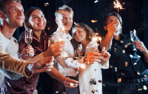 Having fun with sparklers. Confetti is in the air. Group of cheerful friends celebrating new year indoors with drinks in hands