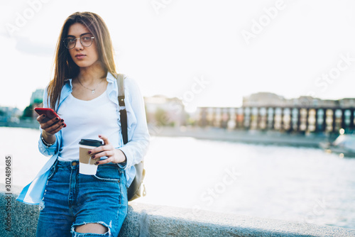 Portrait of attractive brunette traveller in eyewear checking email on modern smarthone standing with coffee in hand outdoors.Female tourist sending sms while looking at camera near publicity area photo