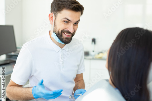 portrait of male smiling dentist talking with client sitting in dental chair