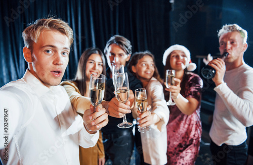 Takes selfie. Group of cheerful friends celebrating new year indoors with drinks in hands