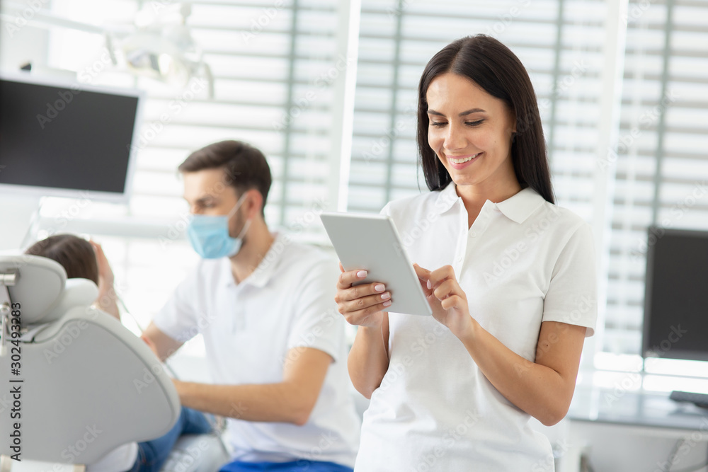 smiling female dentist using digital tablet while her collegue working with small parient on the background smiling female dentist using digital tablet while her collegue working with small parient on the background