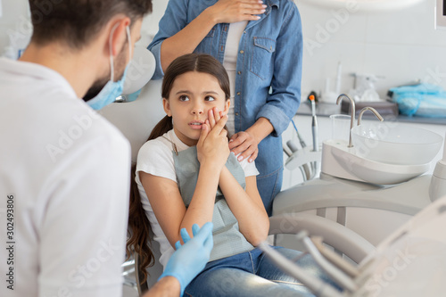 small client with dentist and pain in the teeth sitting in dental chair while mother standing near her for support